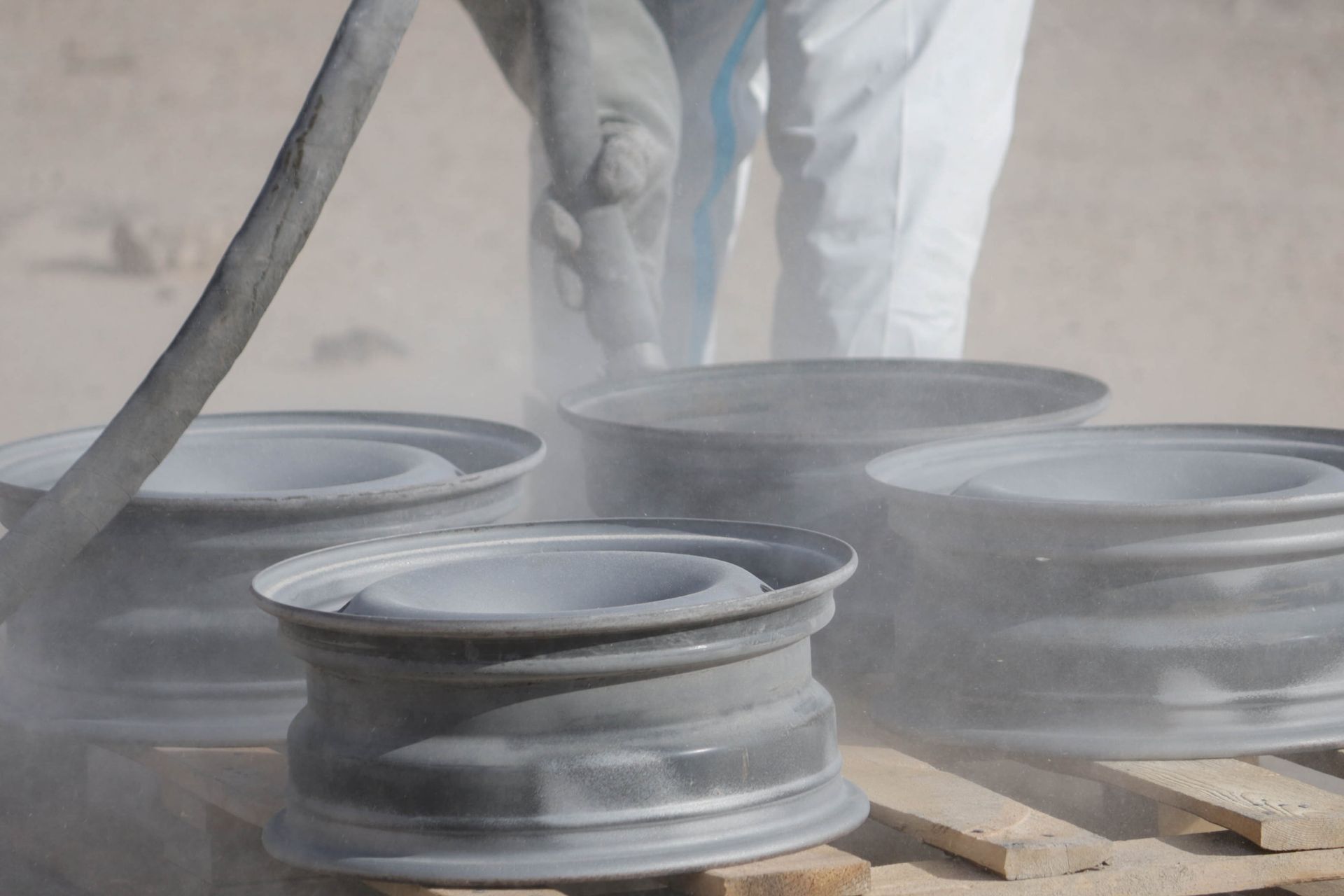 A person wearing protective clothing uses a sandblaster to clean metal wheel rims on a wooden pallet.