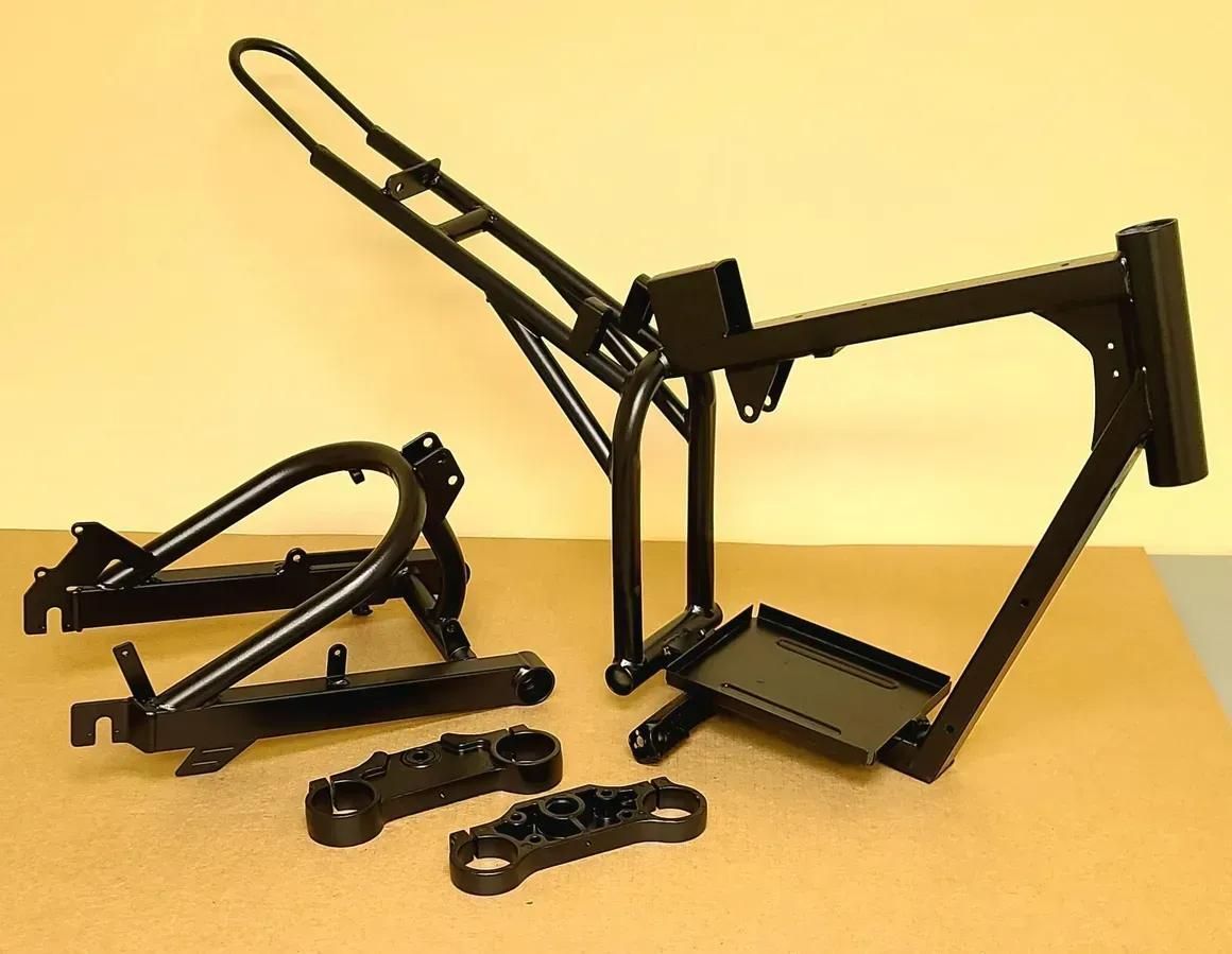 A matte black, disassembled motorcycle frame, swingarm, and triple tree clamps arranged on a flat surface.