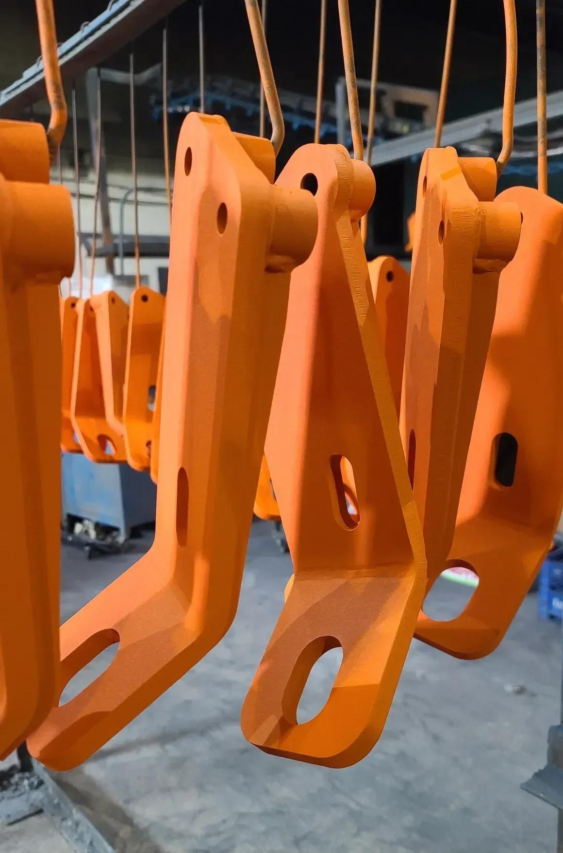 Bright orange metal industrial parts hanging from hooks on a conveyor line in a workshop.