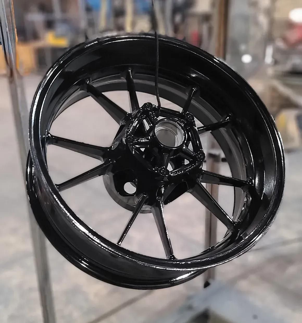 A freshly painted, glossy black motorcycle wheel suspended by a wire in a workshop setting.