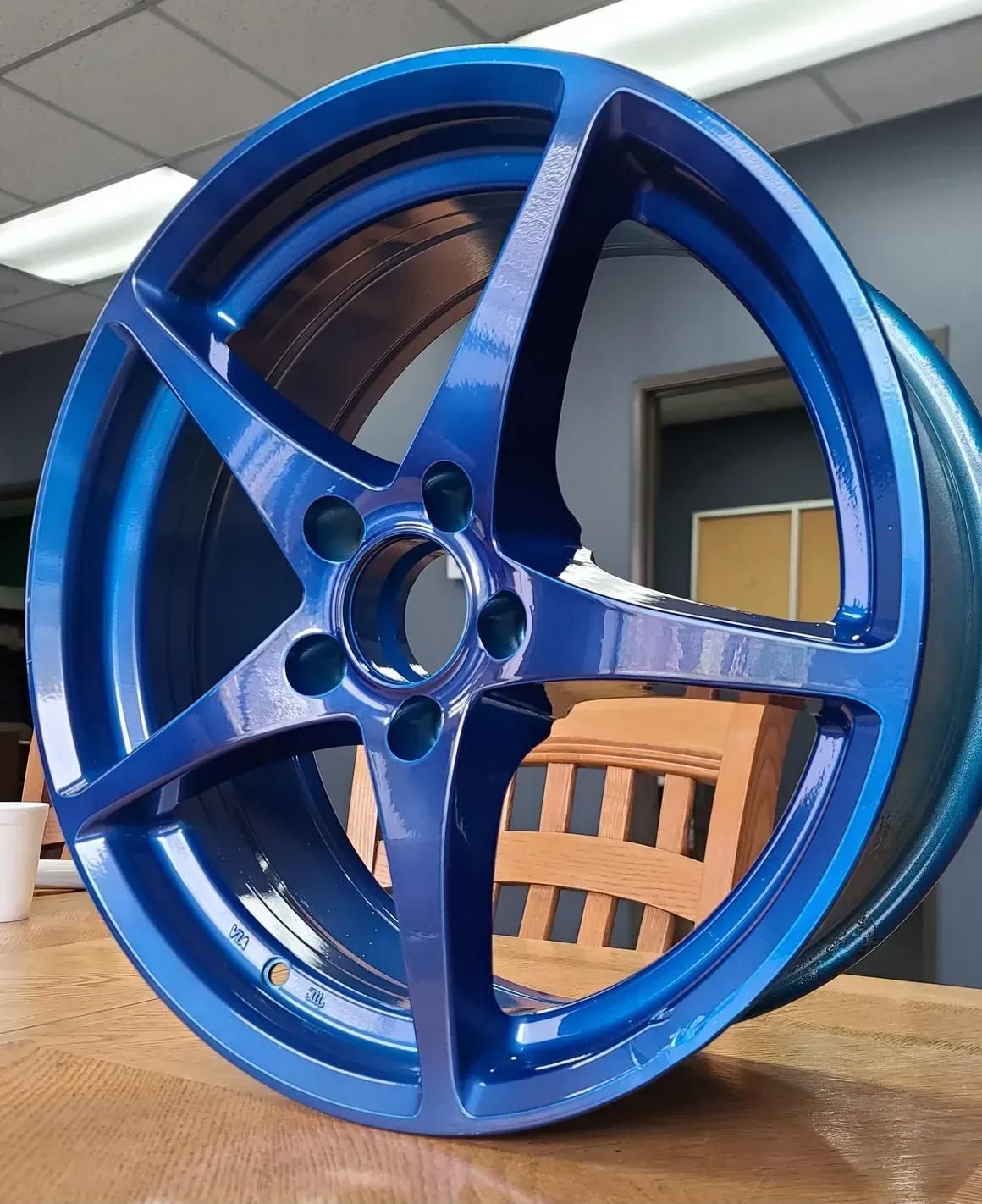 A glossy, vibrant blue five-spoke automotive wheel rests on a wooden table indoors.