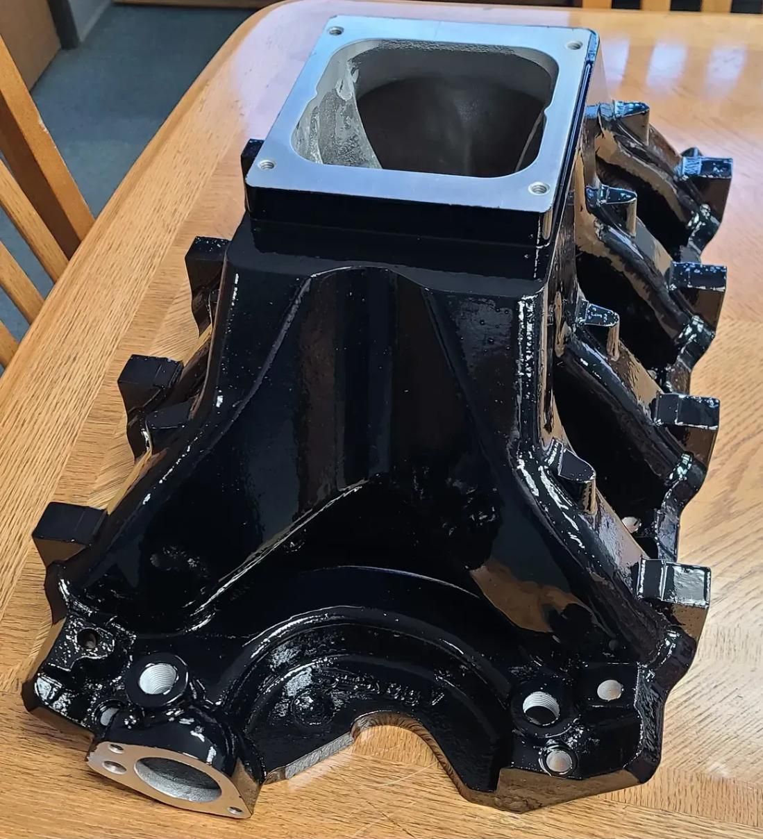 A black painted, high-performance automotive intake manifold sitting on a wooden table.
