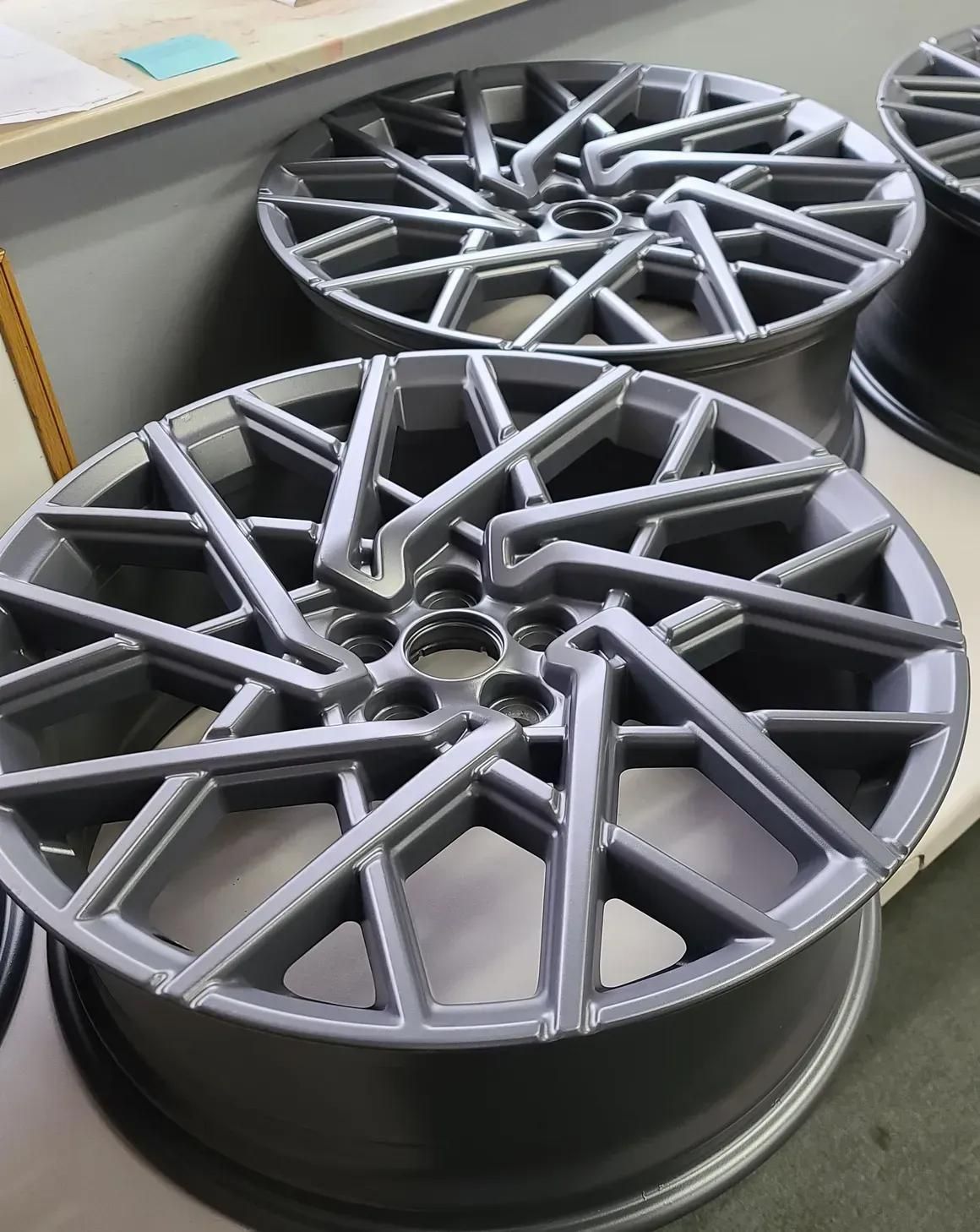 Two matte dark grey, multi-spoke automotive alloy wheels resting on a white surface.