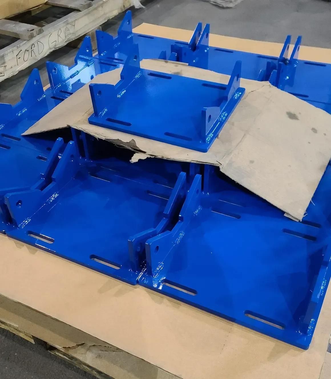 Several bright blue metal mounting brackets with slotted bases stacked on cardboard in an industrial setting.