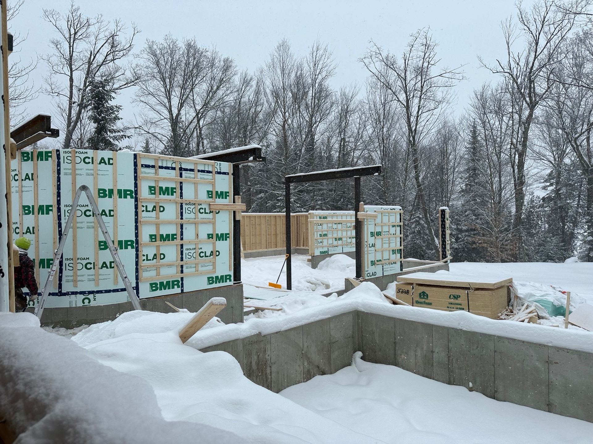 Chantier de construction enneigé avec murs de fondation en béton et matériaux de construction emballés dans une forêt hivernale