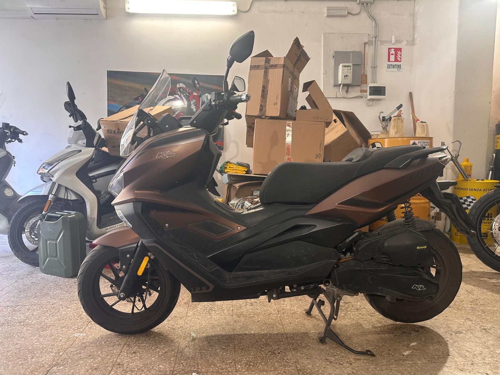 Uno scooter marrone parcheggiato in un garage, con scatole e altre motociclette sullo sfondo.