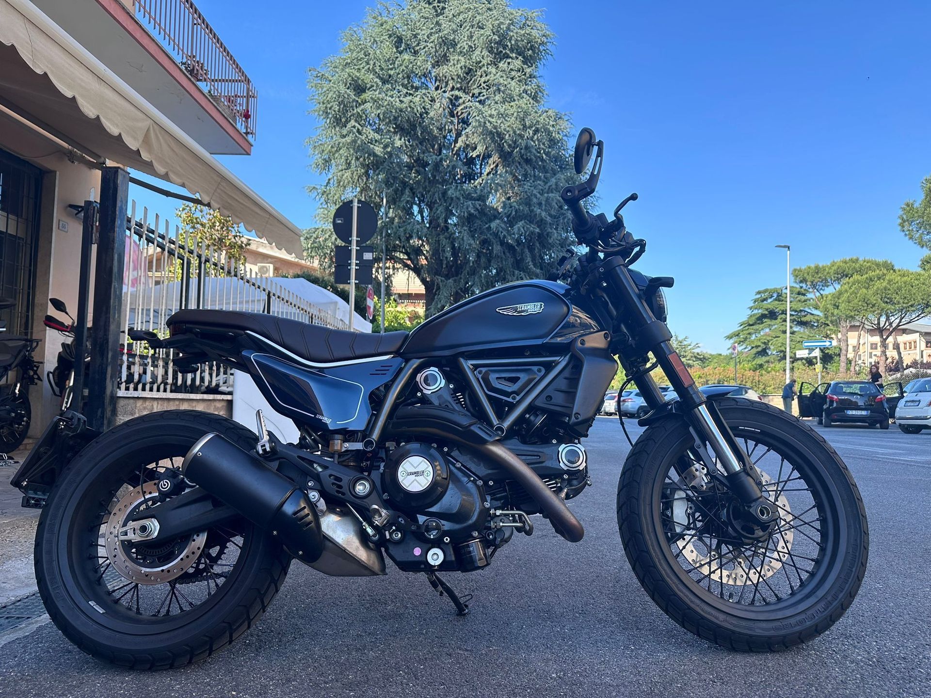 Motocicletta nera parcheggiata su una strada soleggiata accanto a un marciapiede e agli alberi.