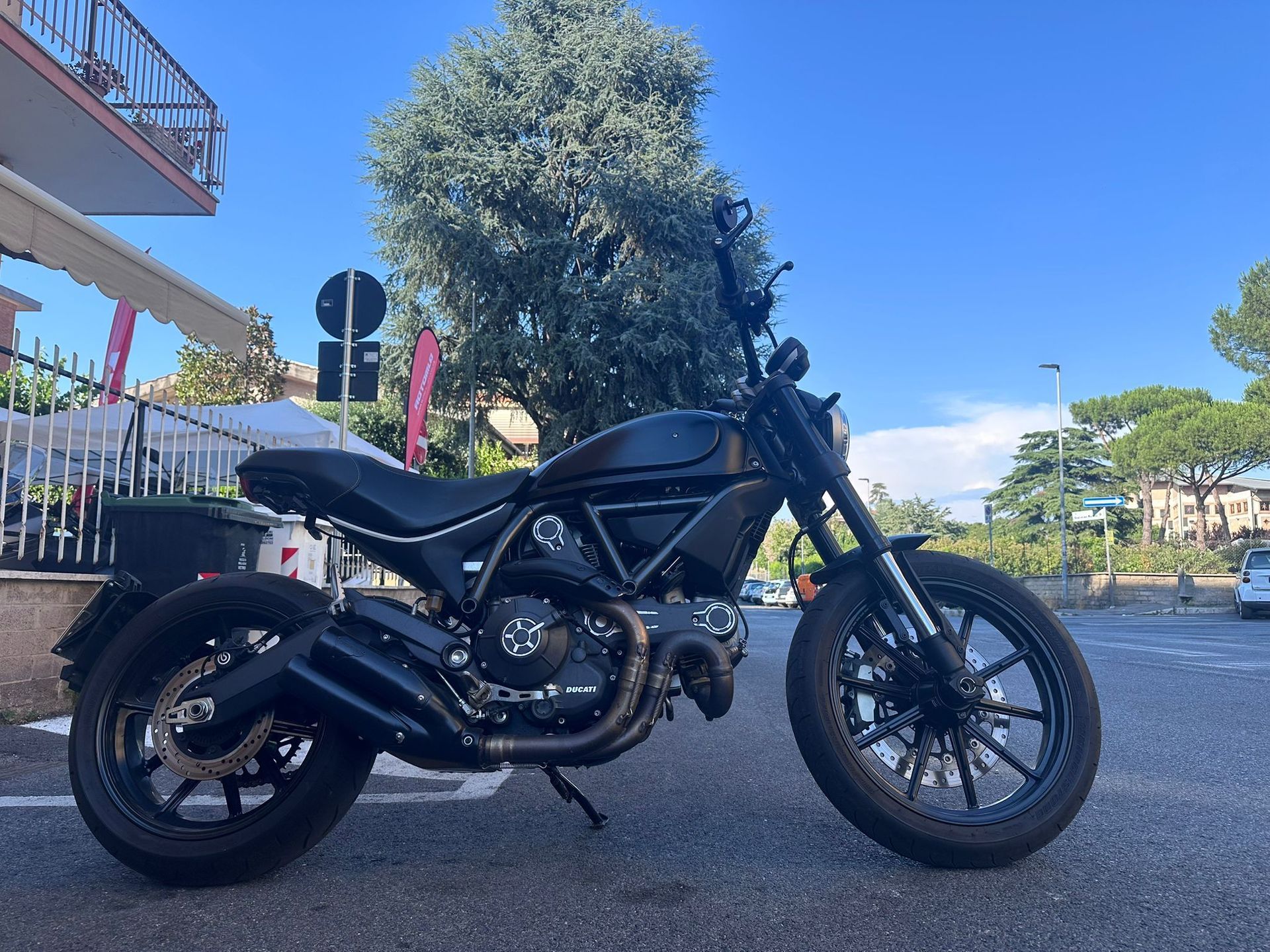 Una motocicletta nera parcheggiata su una strada sotto un cielo azzurro, con alberi ed edifici sullo sfondo.
