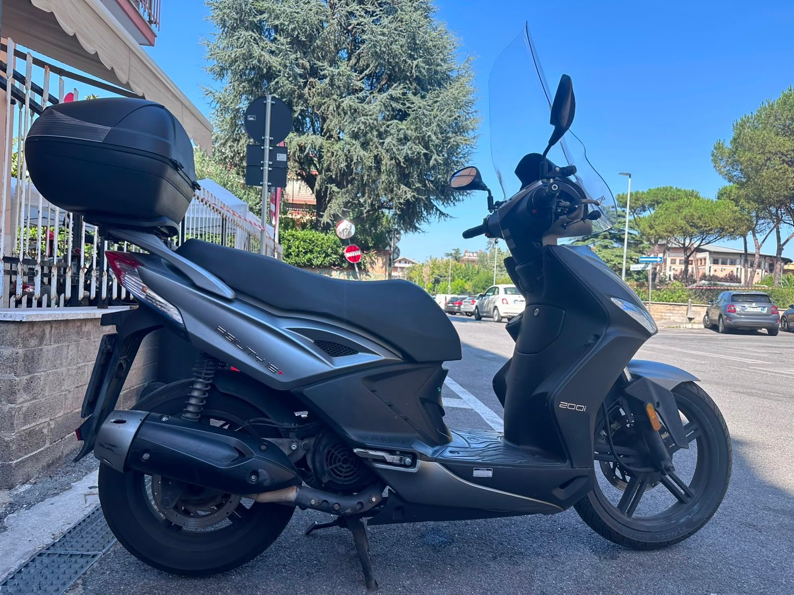 Scooter nero parcheggiato in una strada soleggiata accanto a una recinzione bianca e ad alberi.