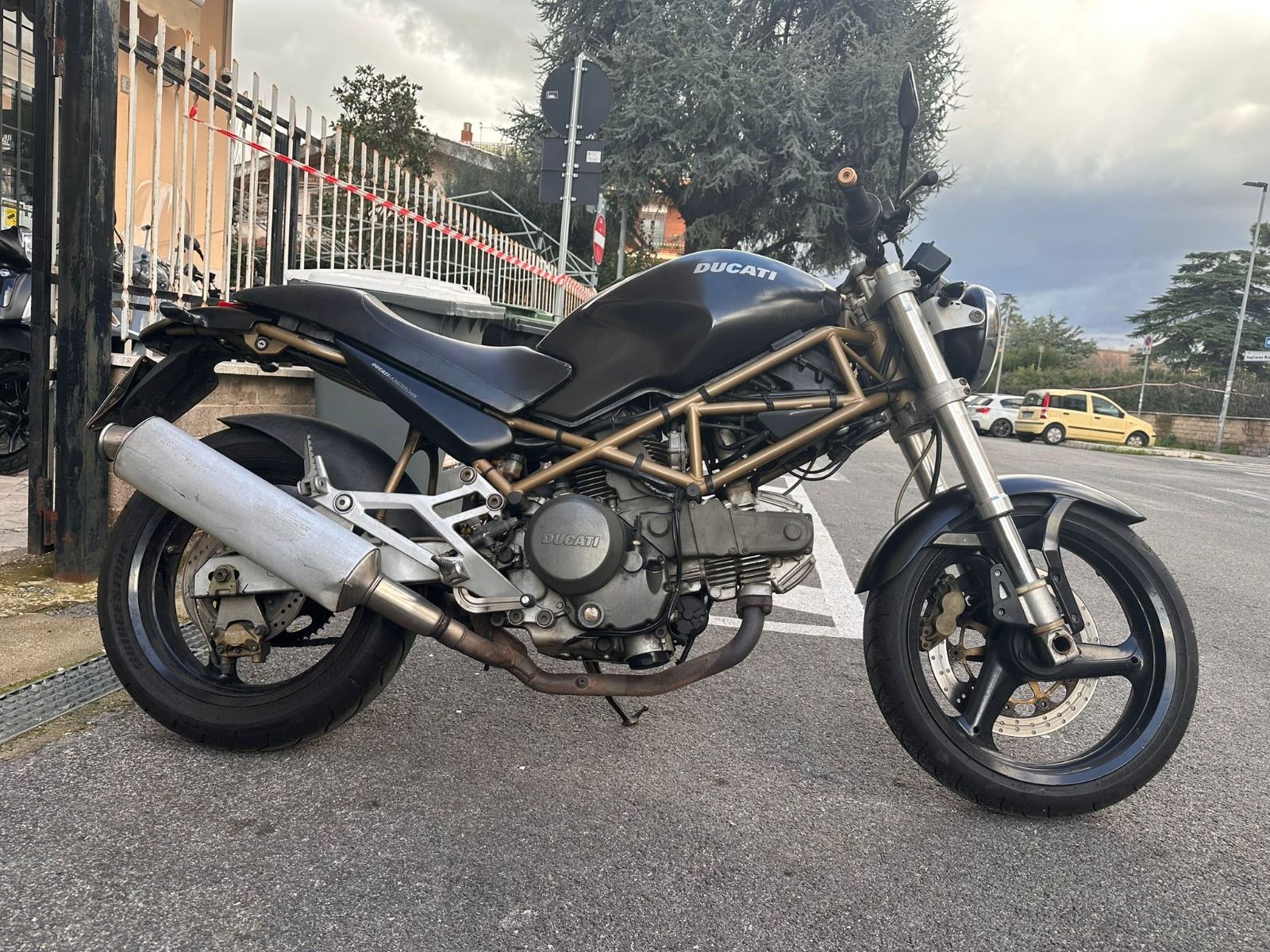 Una motocicletta nera, con telaio a vista e scarico argentato, è parcheggiata in strada accanto a una recinzione metallica.