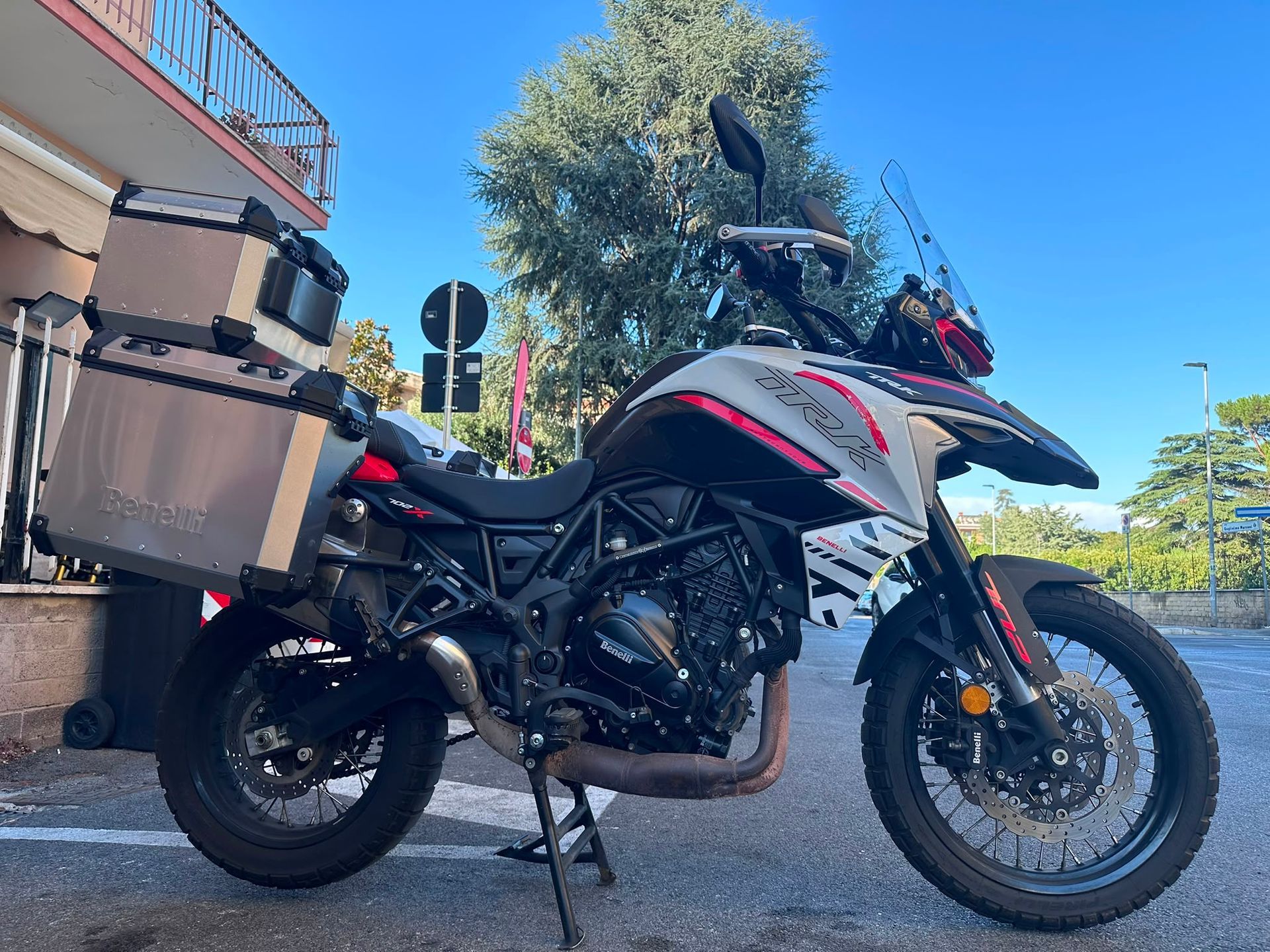 Moto da avventura parcheggiata accanto a un edificio, con borse laterali e parabrezza alto, sotto un cielo azzurro e limpido.