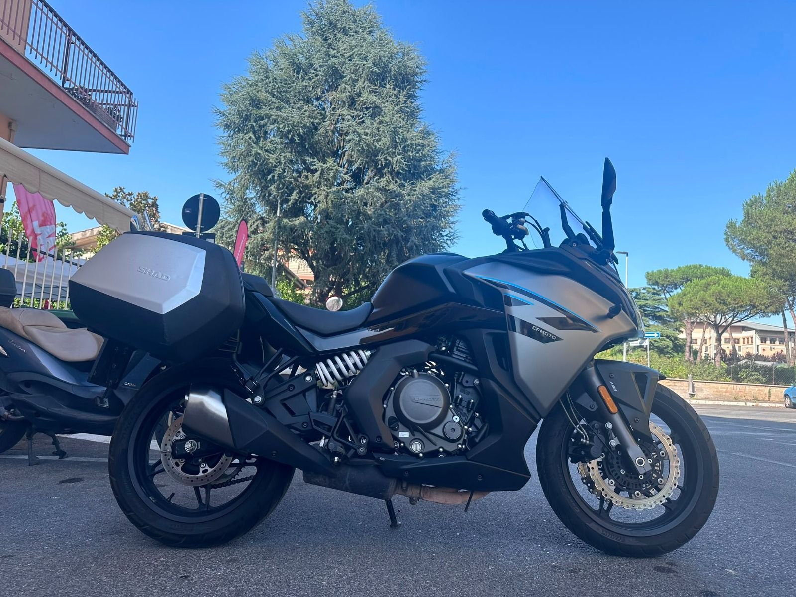 Una motocicletta da turismo nera parcheggiata all'aperto accanto a un albero, sotto un cielo azzurro e limpido.