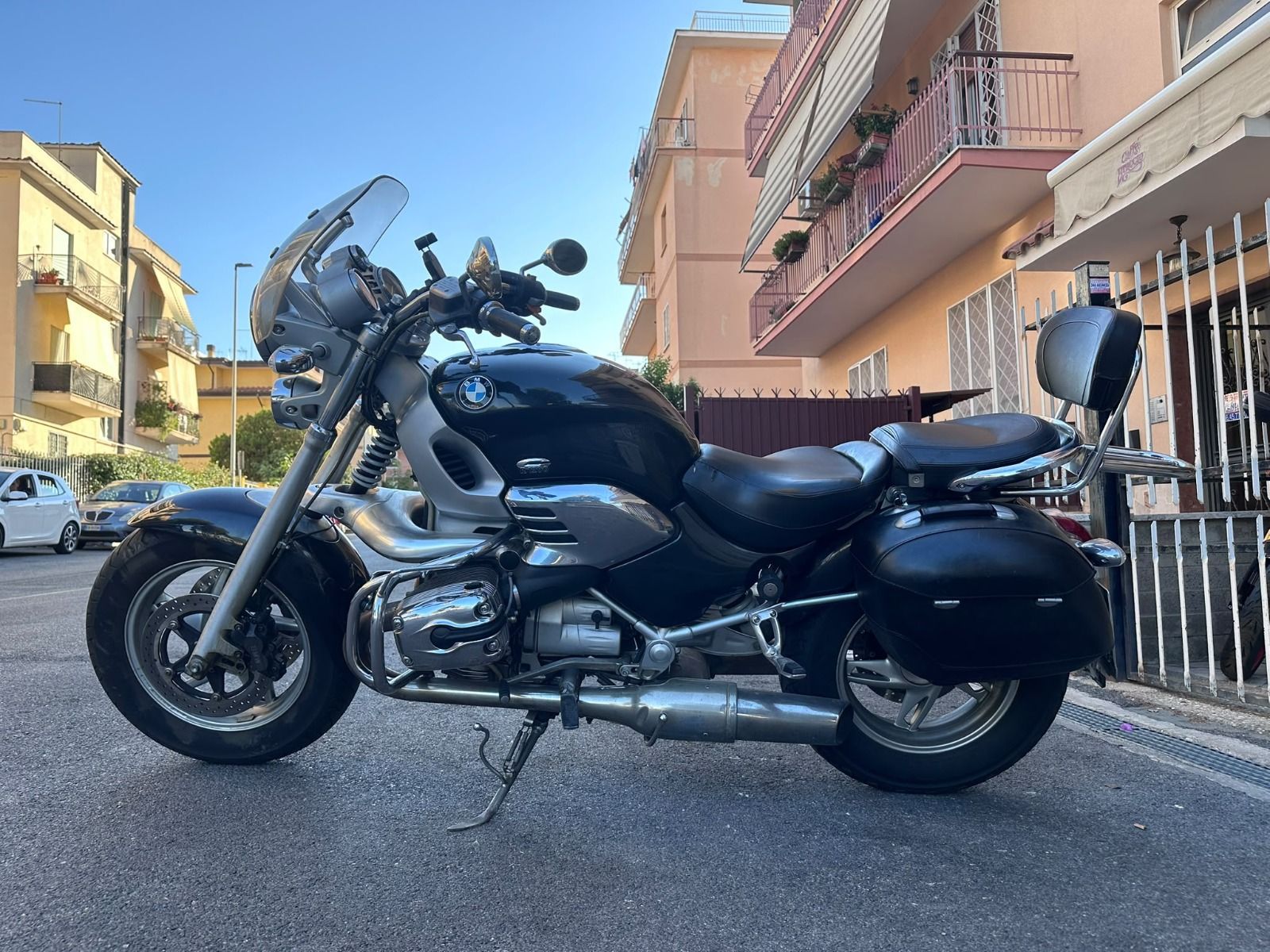 Una motocicletta cruiser nera parcheggiata in una strada soleggiata accanto a dei palazzi residenziali.