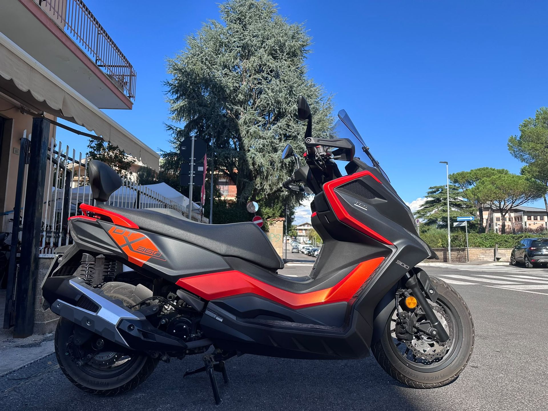 Scooter rosso e nero parcheggiato in una strada soleggiata accanto a un edificio e ad alberi.