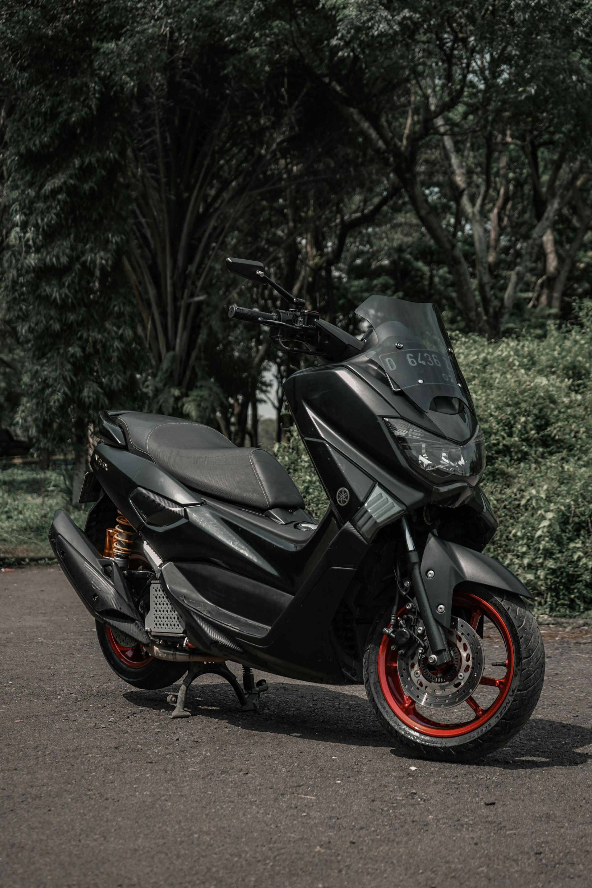 Uno scooter Yamaha NMAX nero opaco con cerchi rossi parcheggiato su un sentiero asfaltato circondato da alberi.