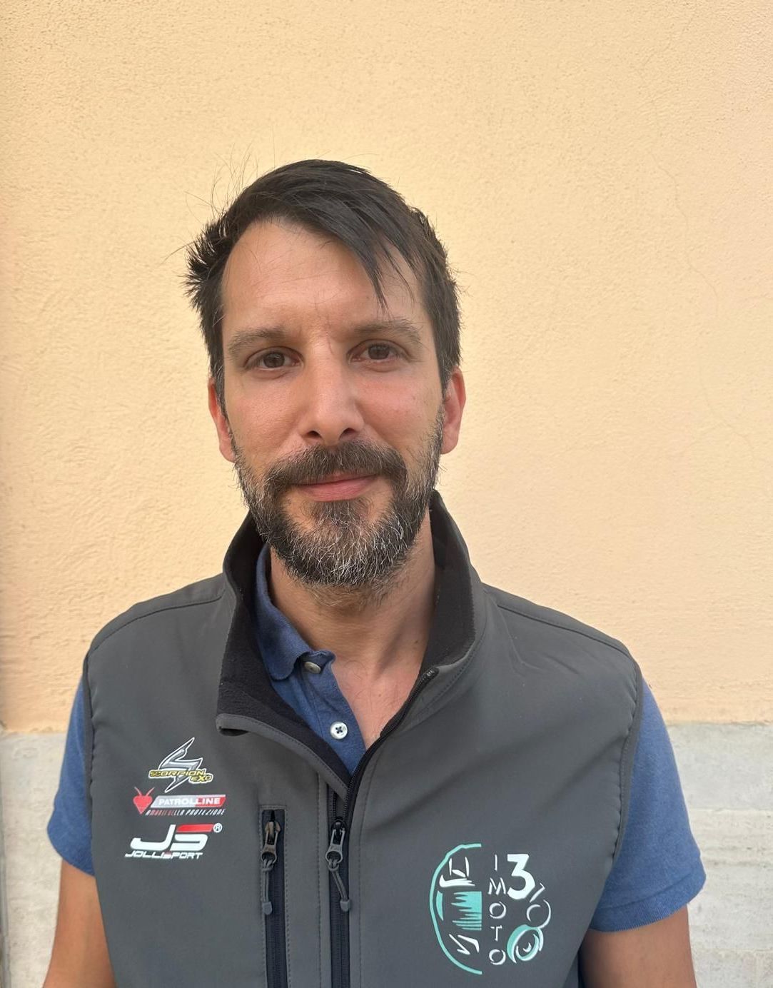 Un uomo con la barba, che indossa una polo blu e un gilet grigio con cerniera e logo, è in piedi davanti a un muro spoglio.