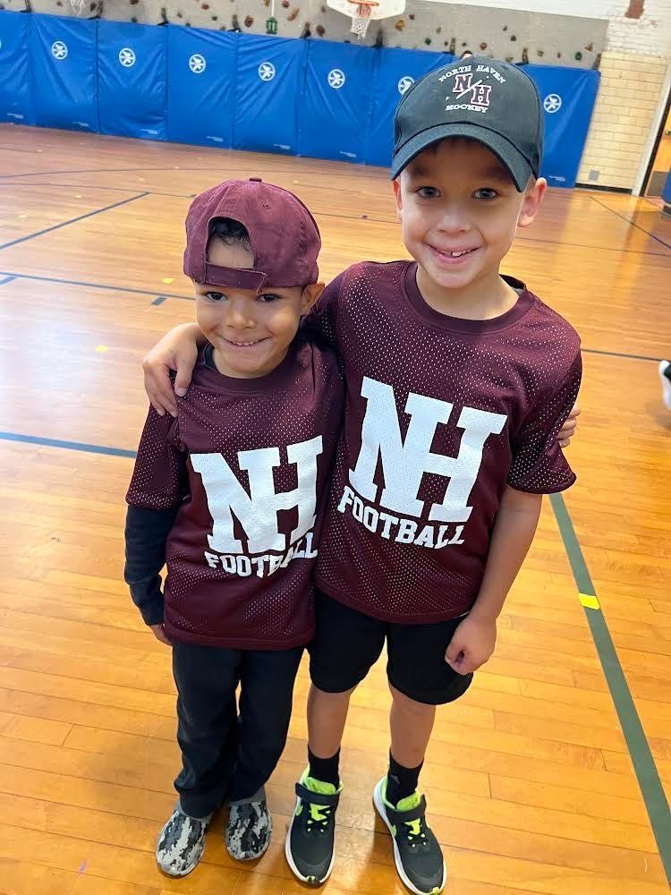 Two young boys wearing nh football shirts are standing next to each other on a gym floor.