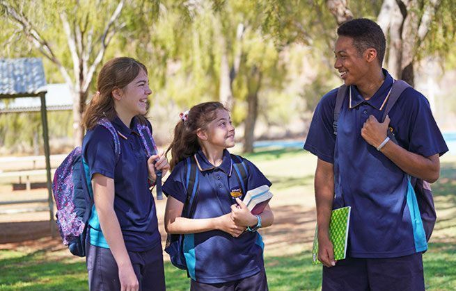 Three students in school uniforms smiling and talking outdoors. One boy and two girls, backpacks on.
