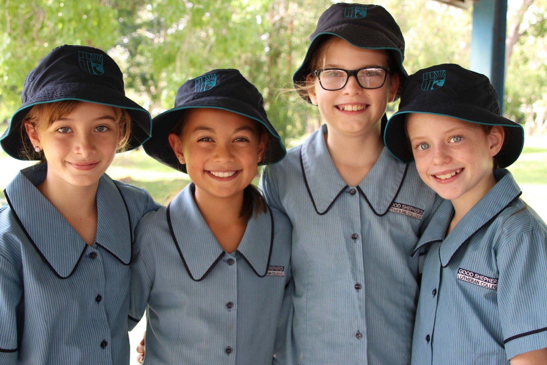 Four smiling schoolgirls in blue uniforms and hats pose outdoors.