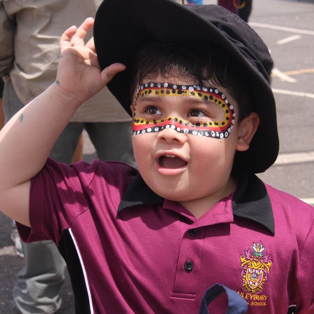 A young child with face paint and a hat, wearing a maroon school uniform, gazes upward with an open mouth.