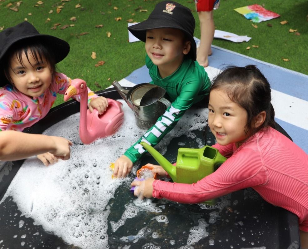 Three smiling children in hats and swimwear play in a soapy water tub outdoors, using watering cans and a flamingo-shaped pourer.