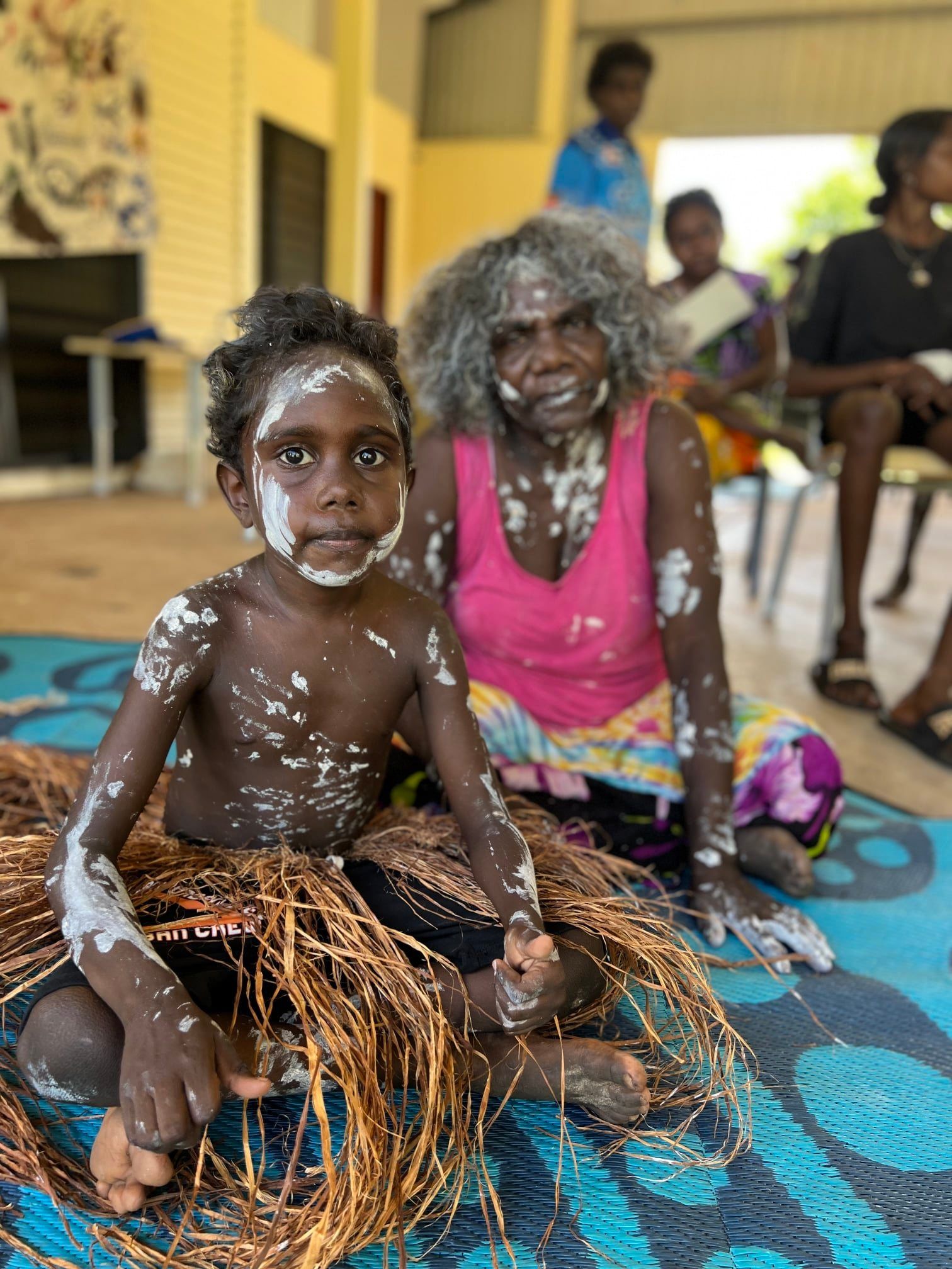 A young child and elder, both with white body paint, sit on a mat with woven material. They appear to be indoors.