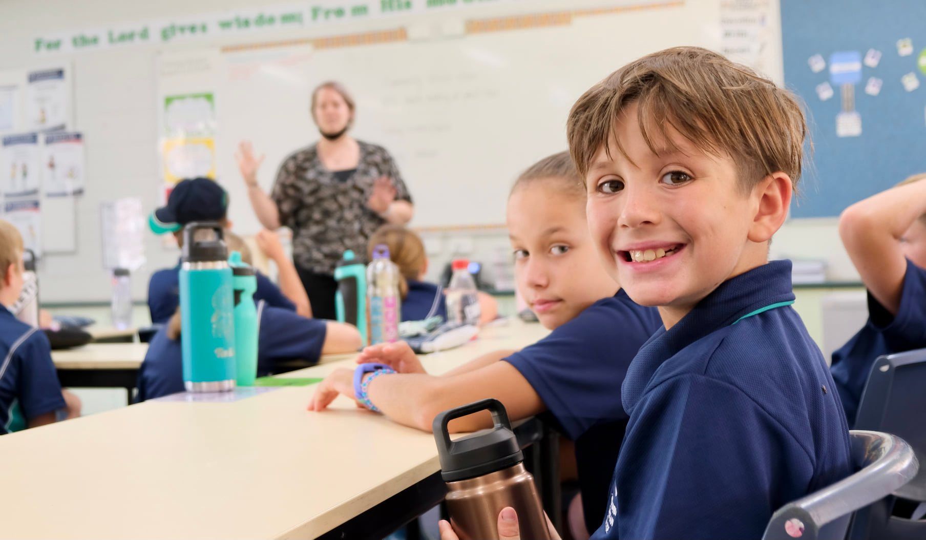Smiling schoolboy in blue uniform at desk, looking at the camera. Teacher in a mask gestures in the background of the classroom.