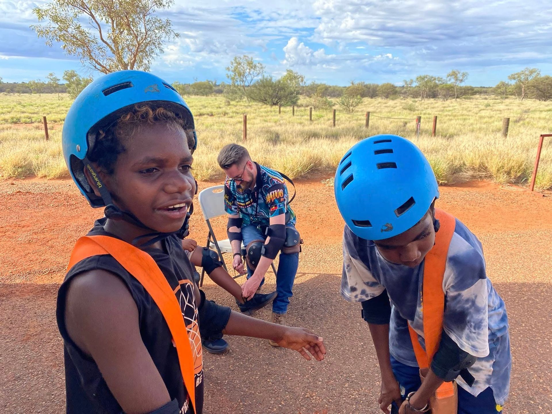 Two young people in blue helmets and orange safety vests on a red dirt road. A person adjusts their gear in the background, the Australian Outback setting is visible.