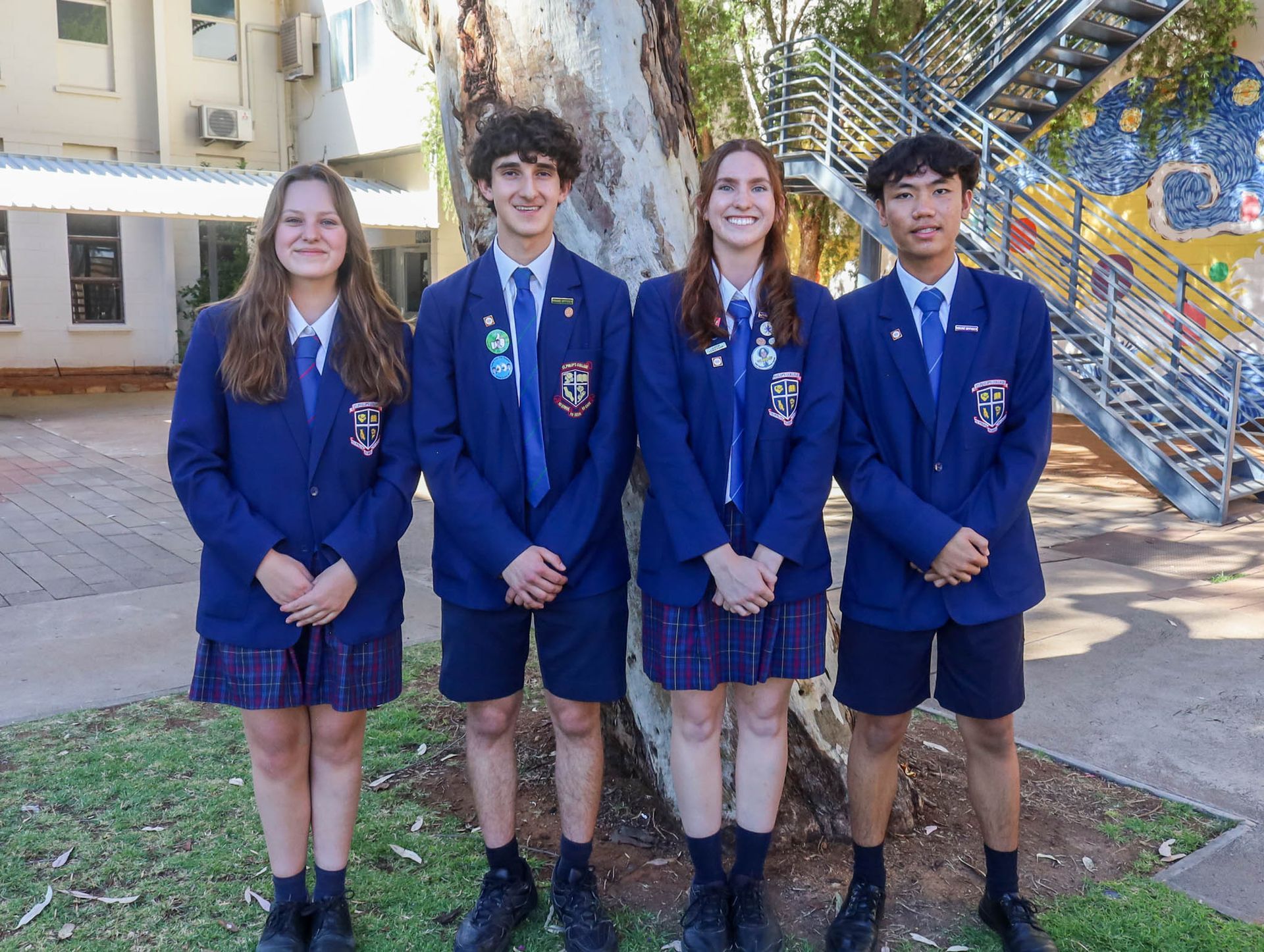 Four students in blue blazers and plaid skirts/shorts stand by a tree outside a school building. They are smiling.