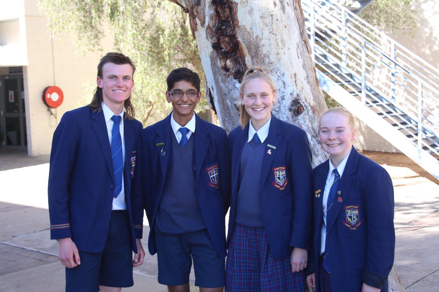 Four high school students in blue blazers stand outdoors smiling, near a tree and stairs.