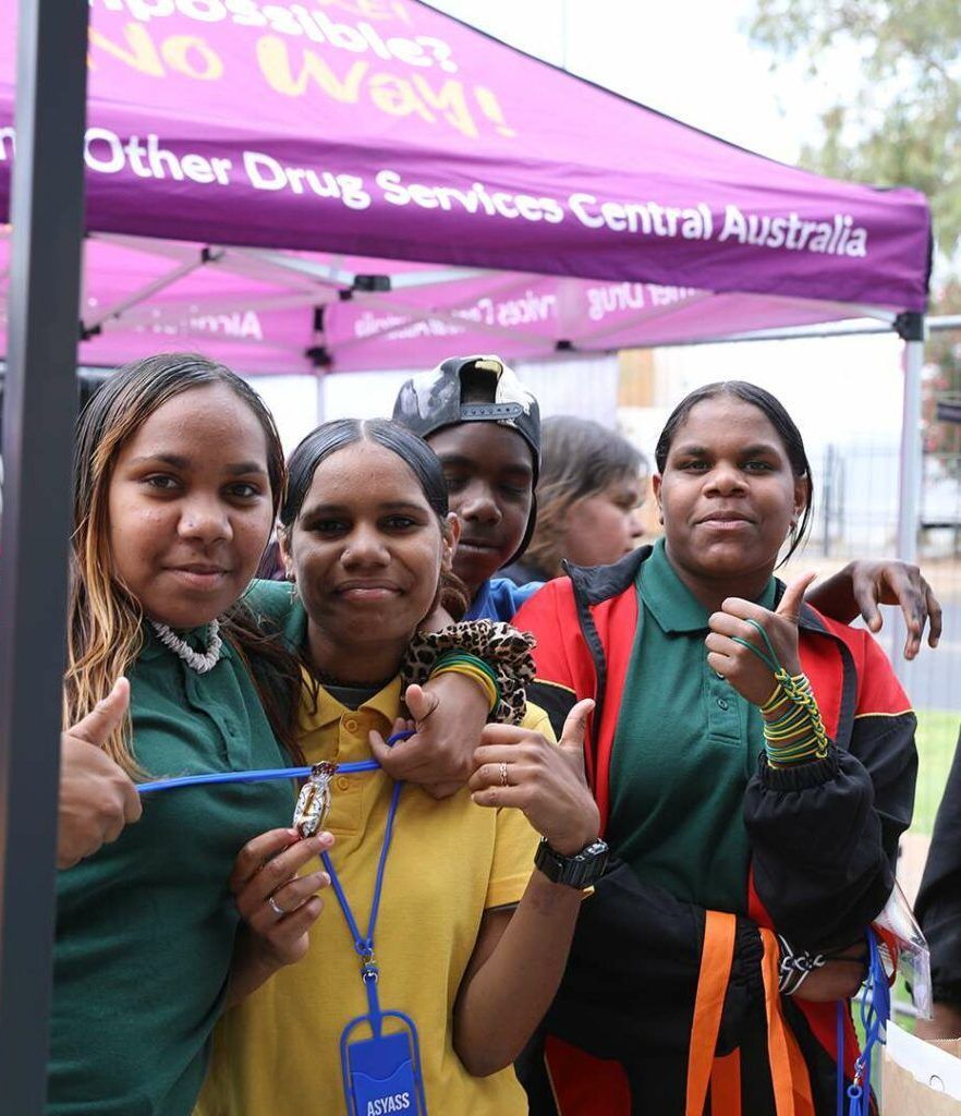 Group of smiling young people in front of a purple tent with text, giving thumbs up.