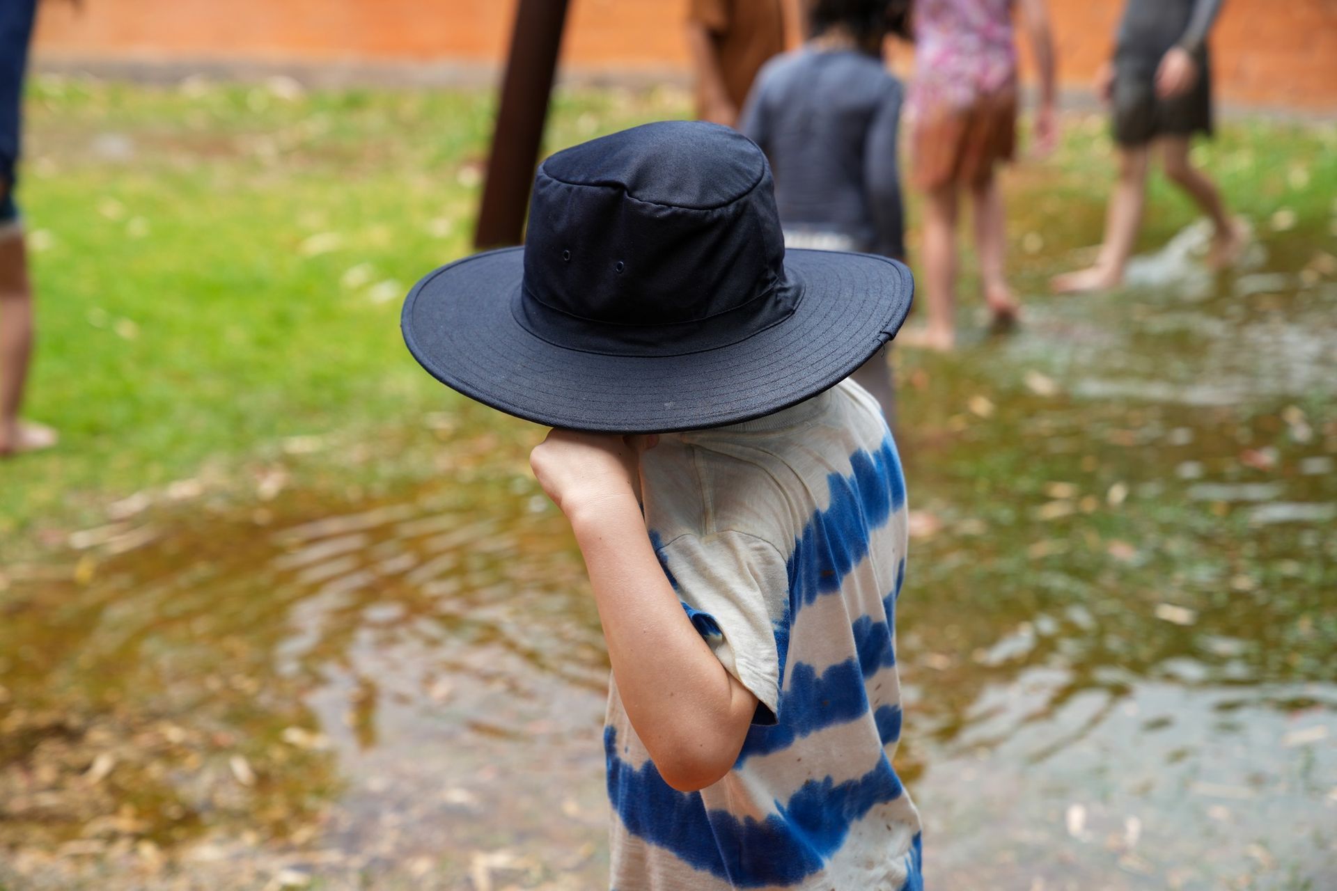 Child wearing a large blue hat, standing in a puddle, partially obscured. Others in the background.