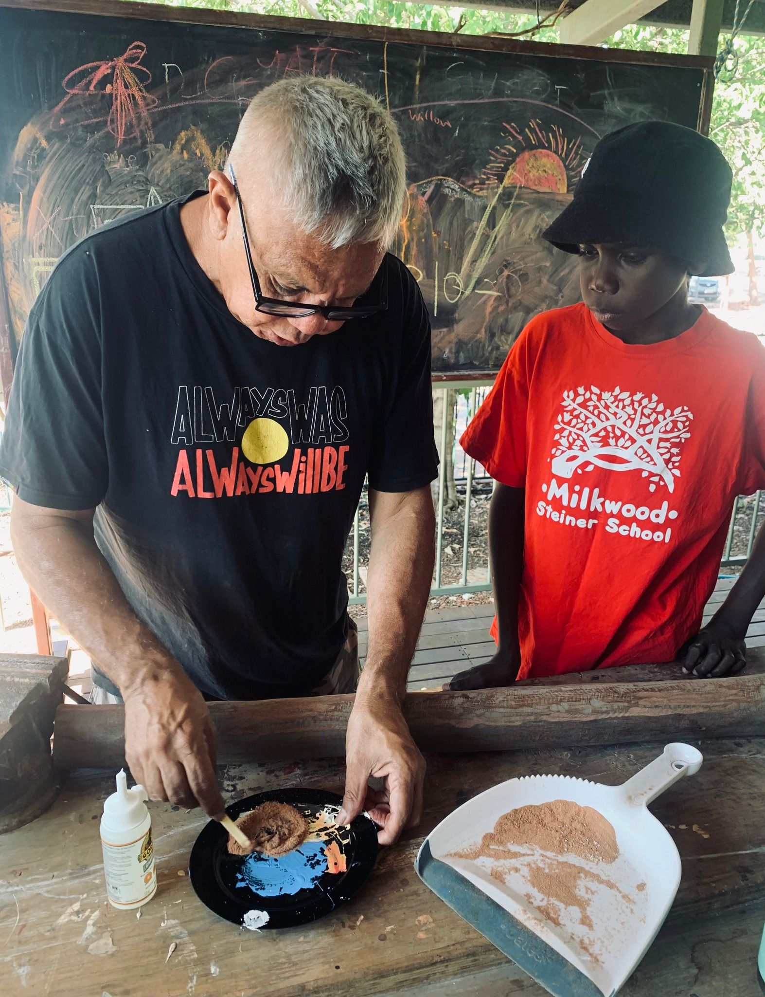 Man with grey hair and glasses working on a craft project with a child wearing a red t-shirt and bucket hat outdoors at a wooden table.