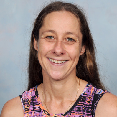 Smiling woman with long brown hair, wearing a patterned purple and orange top, against a light blue background.