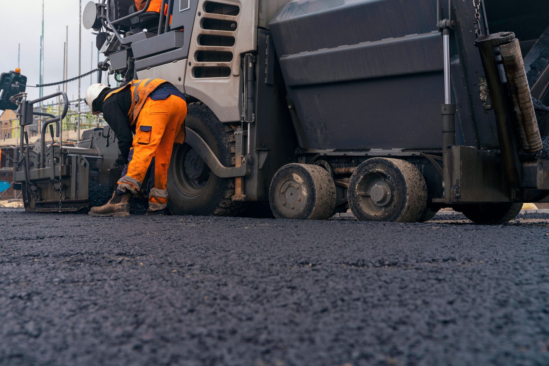 An asphalt paving company worker in orange safety gear checks a paver on freshly laid dark asphalt. An asphalt paving company worker in orange safety gear checks a paver on freshly laid dark asphalt.