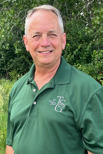 Smiling man in green polo shirt standing outdoors by tall grass and trees