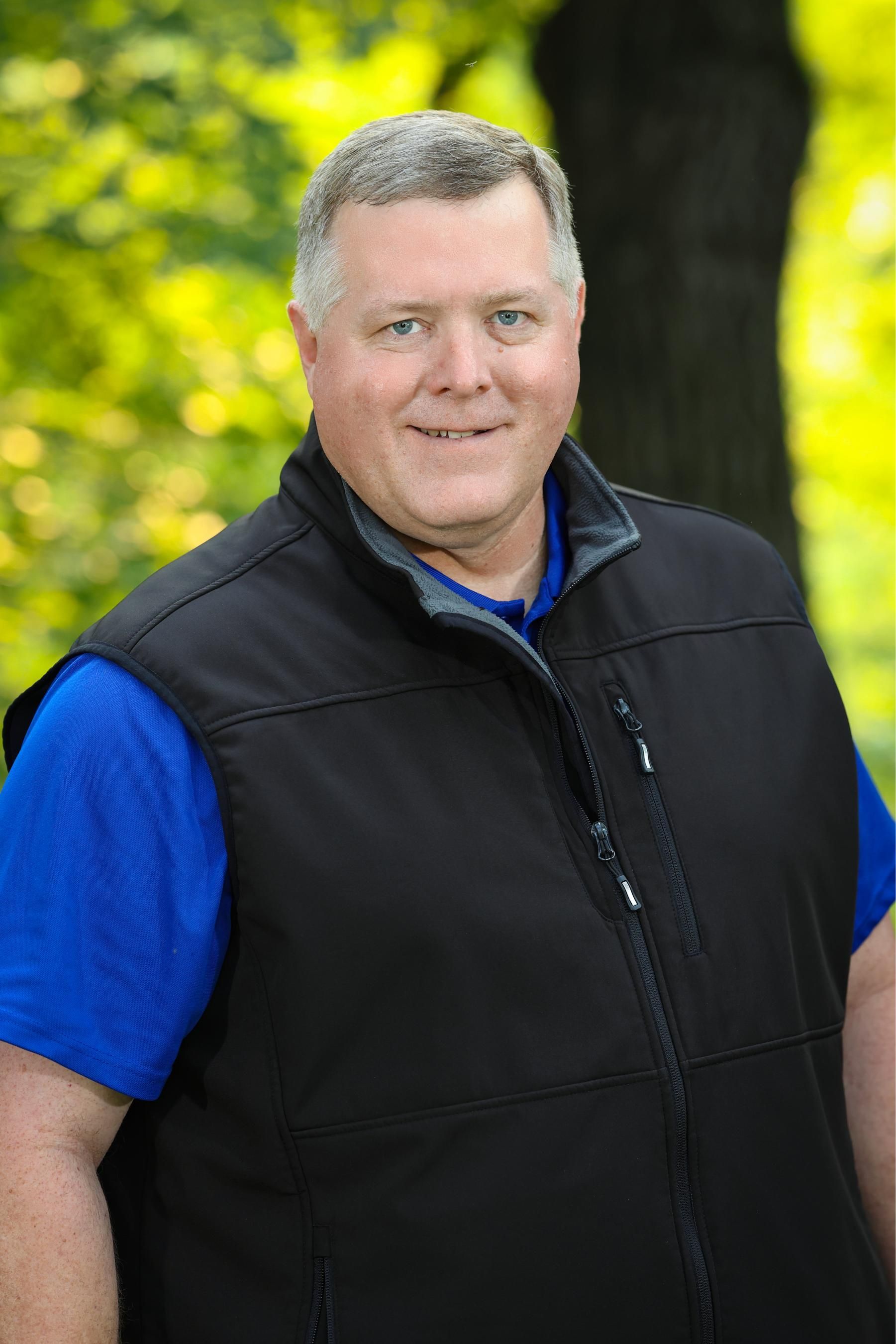 A man wearing a black vest and a blue shirt is standing in front of a tree.