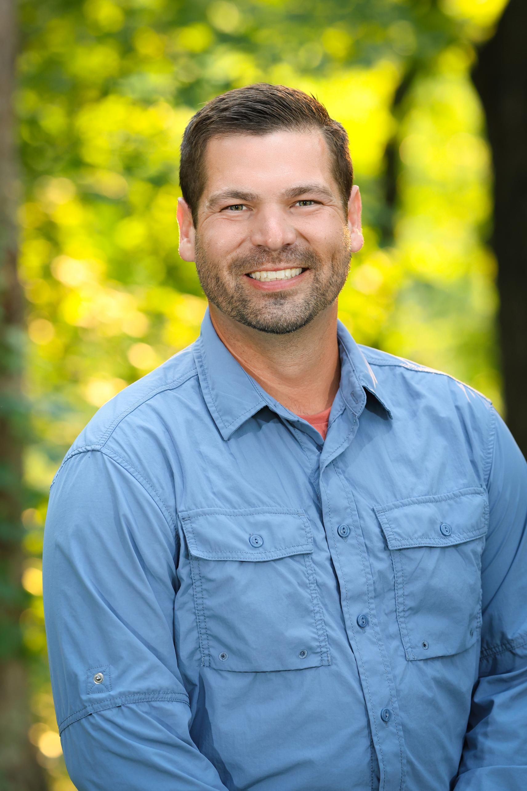 A man in a blue shirt is smiling for the camera while standing in front of trees.