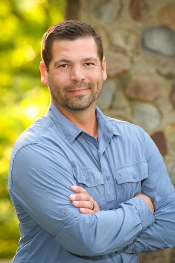 A man in a blue shirt is standing with his arms crossed in front of a stone wall.