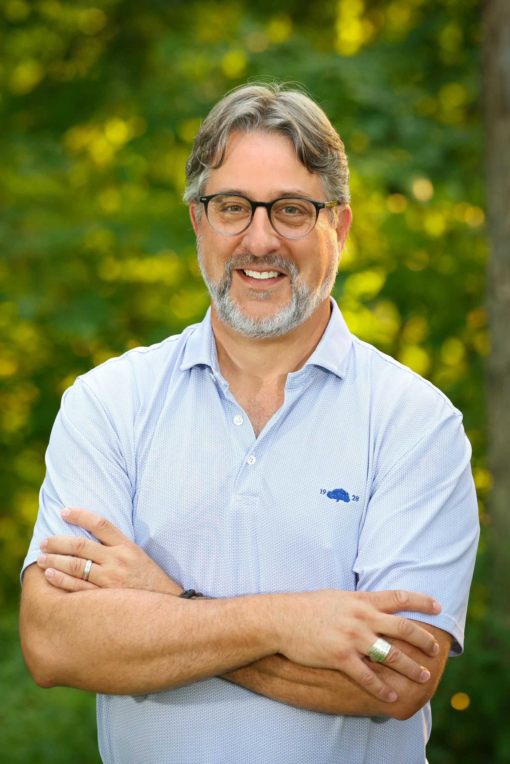 A man with glasses and a beard is standing with his arms crossed and smiling.