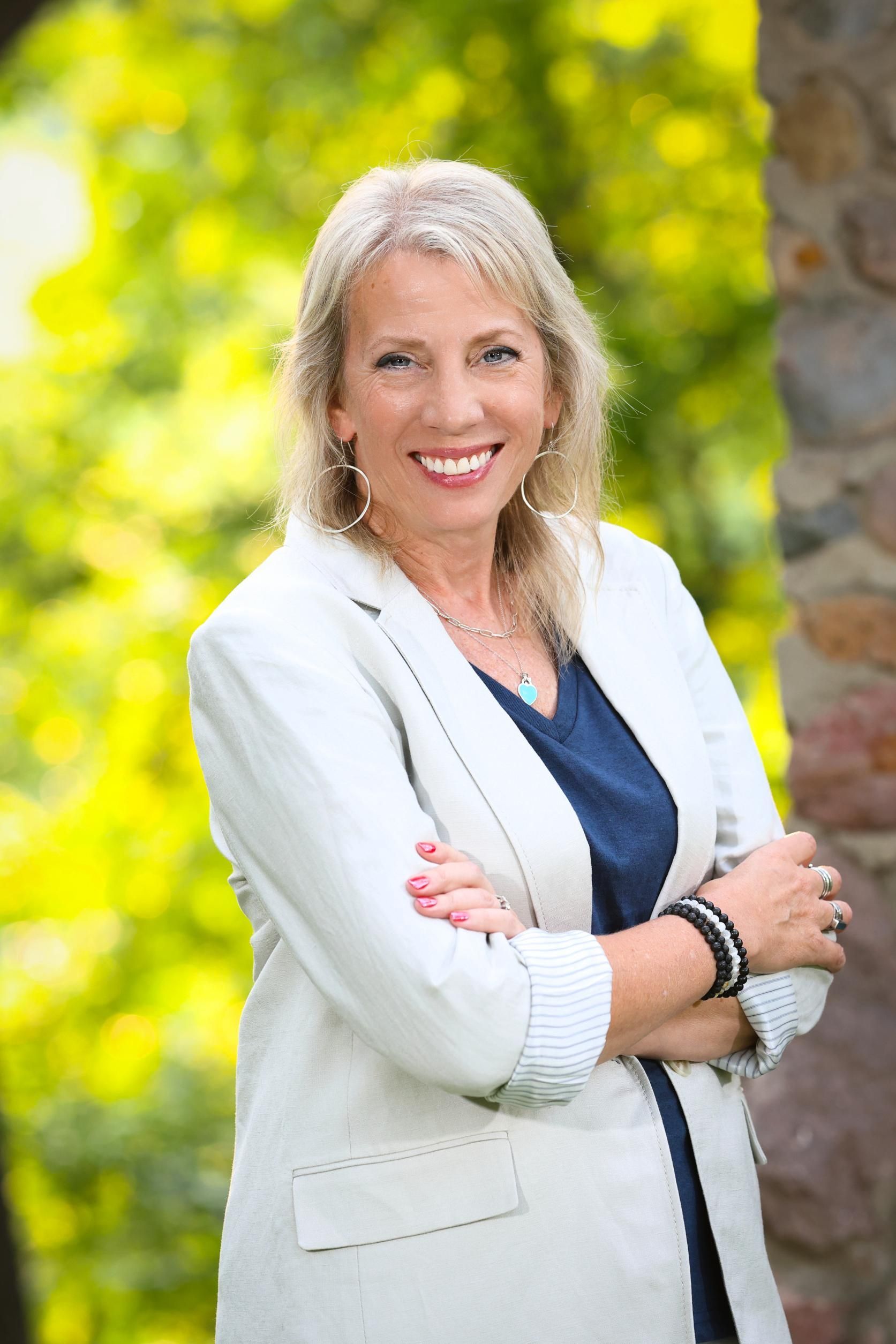 A woman in a white jacket and blue shirt is standing with her arms crossed and smiling.