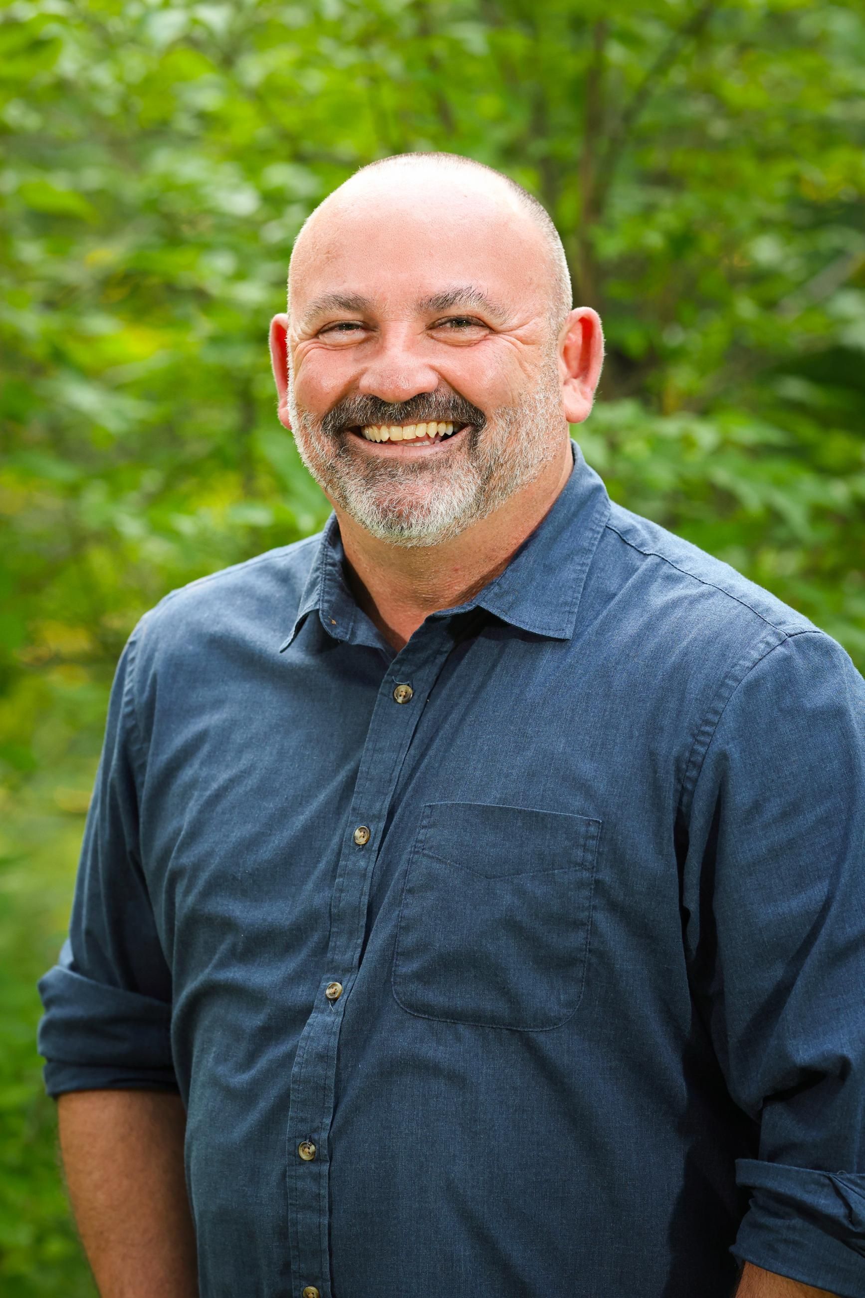 A bald man with a beard is smiling for the camera while wearing a blue shirt.