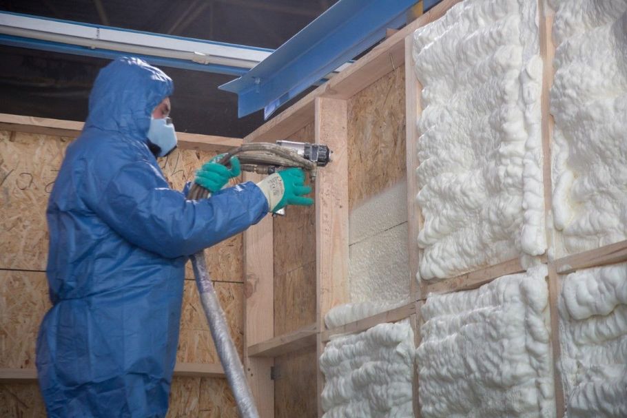 Worker in blue protective suit sprays insulation foam inside a wall cavity.