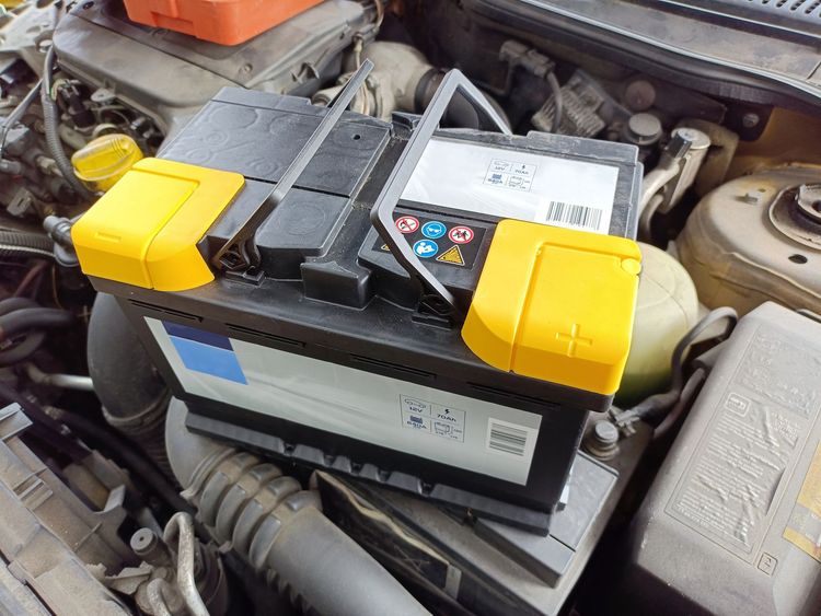 Batteria per auto nel vano motore, con copri terminali gialli e una cinghia nera che attraversa la parte superiore.