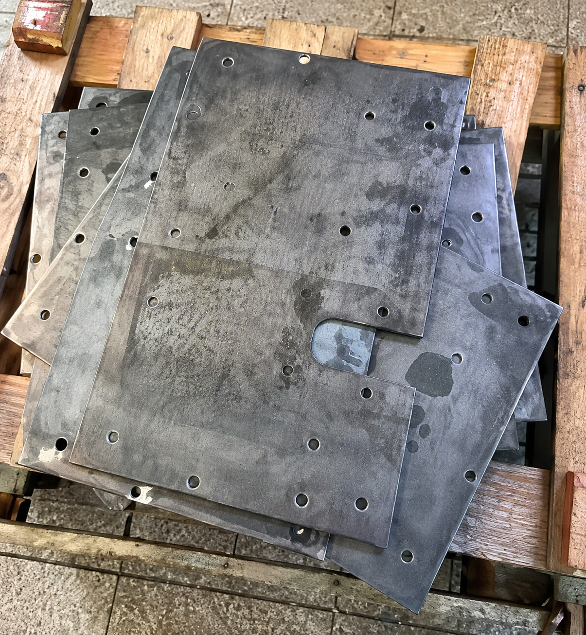 Piastre metalliche grigie impilate, con fori per bulloni, appoggiate su un pallet di legno all'aperto.