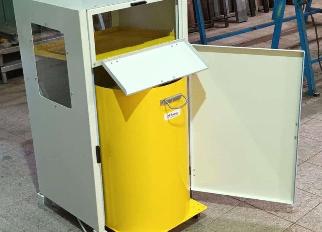 Contenitore giallo per la raccolta differenziata con sportello laterale bianco aperto, in un'area industriale interna.