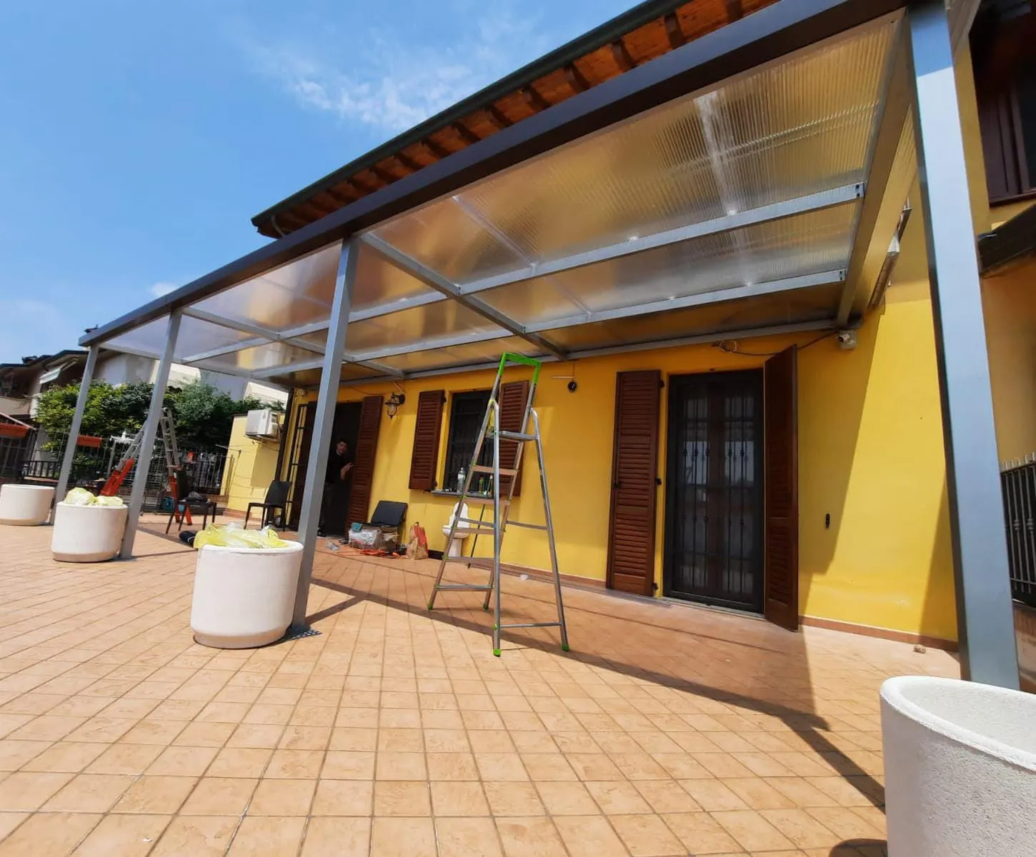 Patio di una casa gialla con tenda da sole in metallo, cortile piastrellato e scala appoggiata al muro.