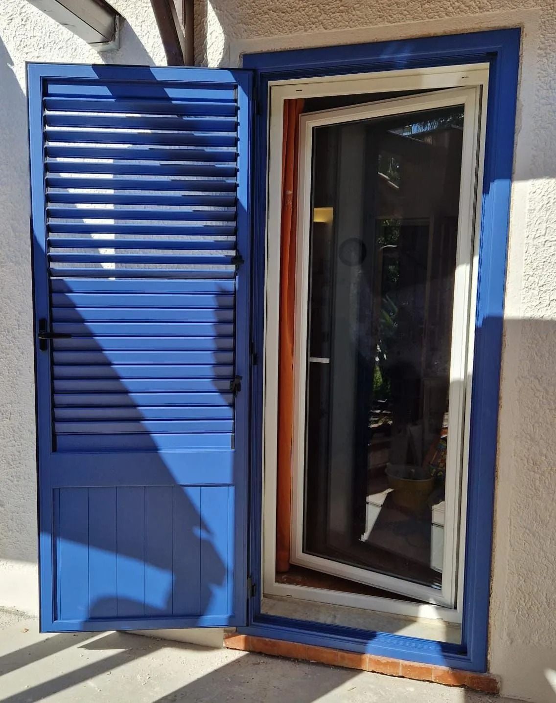 Persiana blu accanto a una porta a vetri con cornice bianca in una parete esterna intonacata.