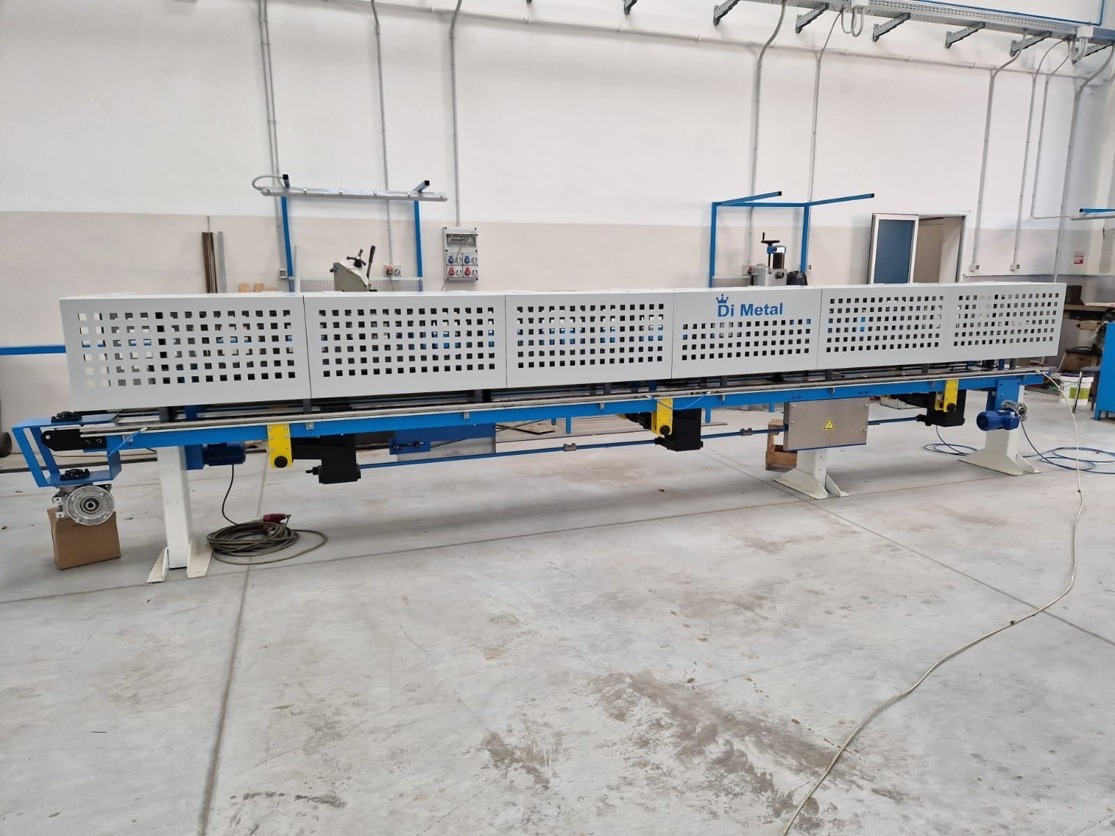 Lungo macchinario industriale su supporti blu in un'officina con un pannello metallico perforato.