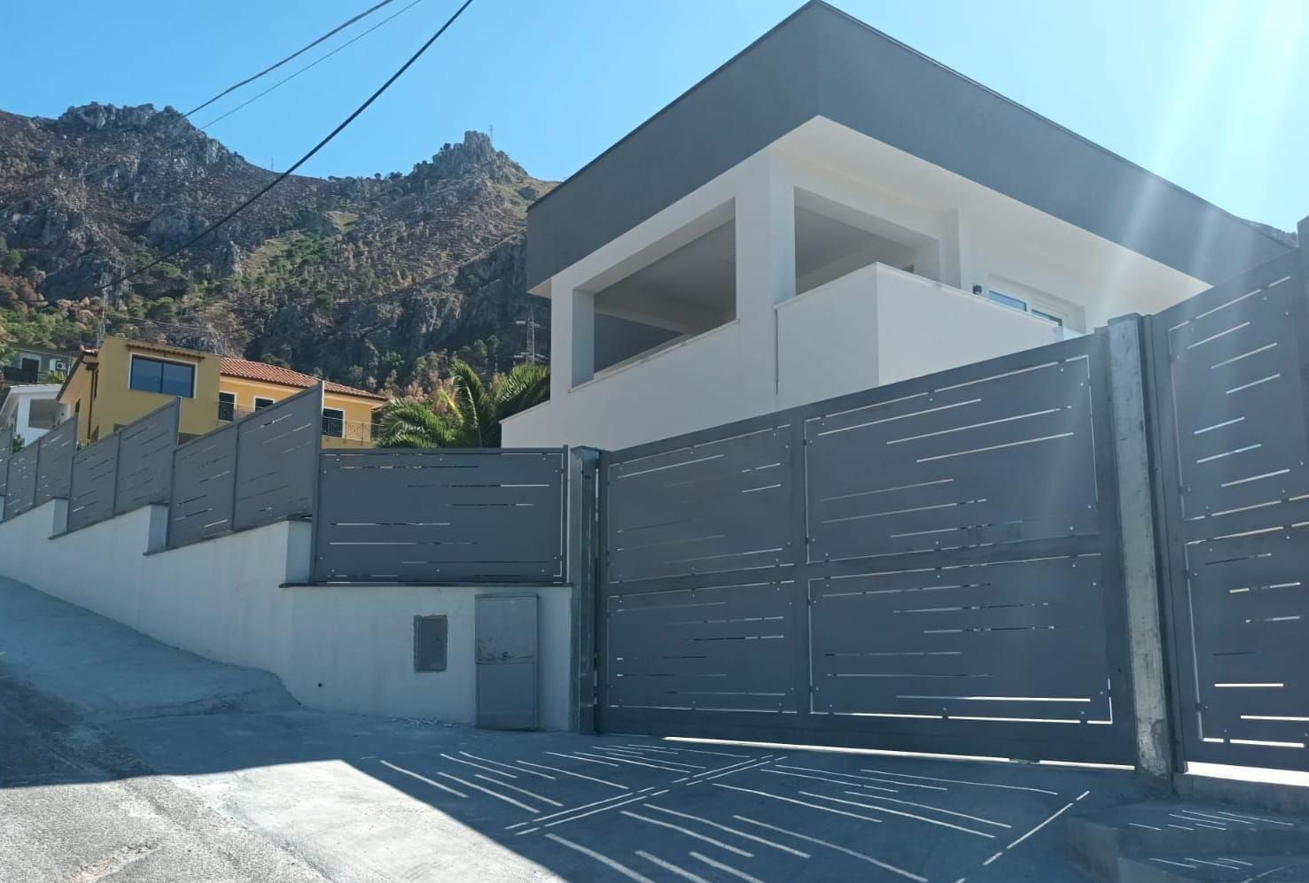 Casa moderna bianca con cancello grigio su una strada soleggiata in collina, con le montagne sullo sfondo.